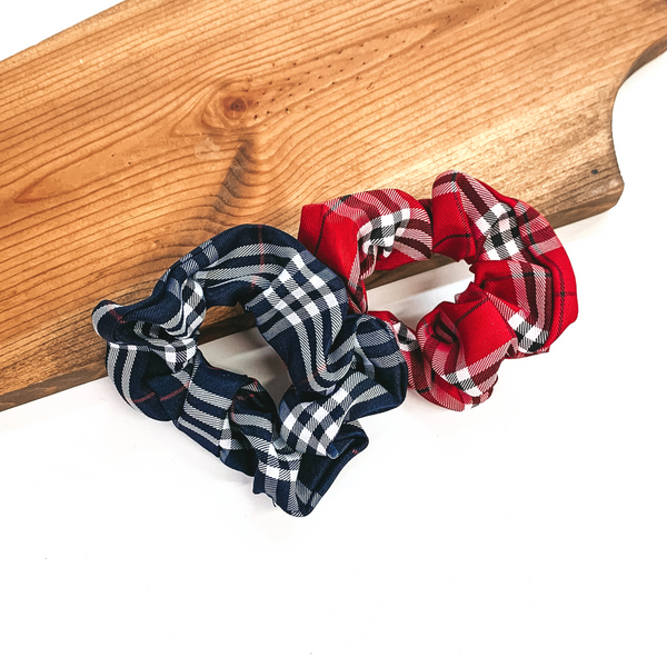These are two plaid print scrunchies, the left one is navy blue and right  one is in red. Both of these scrunchies are taken leaning on a brown  necklace board and on a white background.