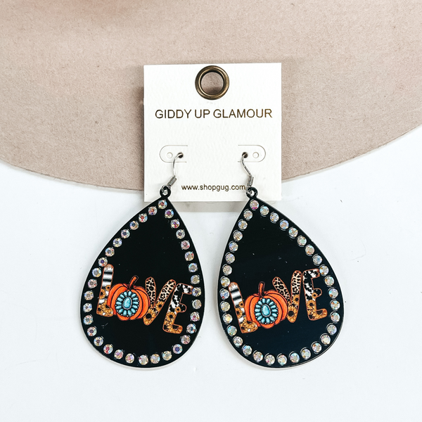 These are teardrop earrings in black with AB crystals all around. In the middle it says 'Love' , each letter has different prints and designs. The 'O' is an orange pumpkin with a turquoise concho in the center. These earrings are taken laying on a beige color hat brim and on a white background.