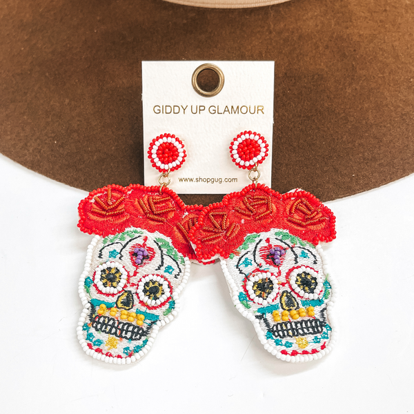 These are sugar skull post earrings in white and other colors. There are red  flowers in the top with beads and stitching. The skull part has white beads  around the eyes, yellow beads in the mouth, and black beads in the eyes.  The rest has multicolored stitching all around. These earrings are taken on  a dark brown hat brim and on a white background.