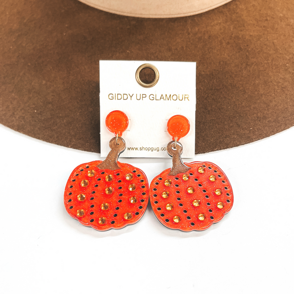 Pictured on a white and brown background are acrylic, orange glitter pumpkin earrings. These earrings include holes as and crystals as detailing.