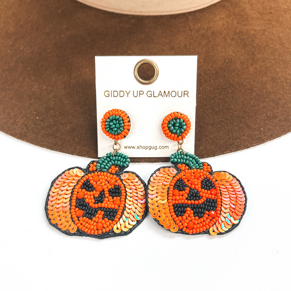 Green and orange circle beaded post back earrings with a hanging beaded pendant. This beaded pendant is shaped like a pumpkin with a Jack O' lantern face. These earrings are pictured on a white and brown background. 