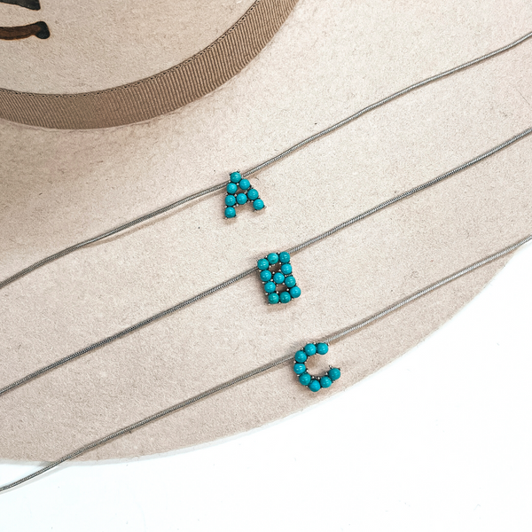 These are three turquoise stone initial necklaces with a thin, silver  snake chain. From top to bottom; A, B, C. These necklaces are taken  laying on a beige felt hat and on a white background.
