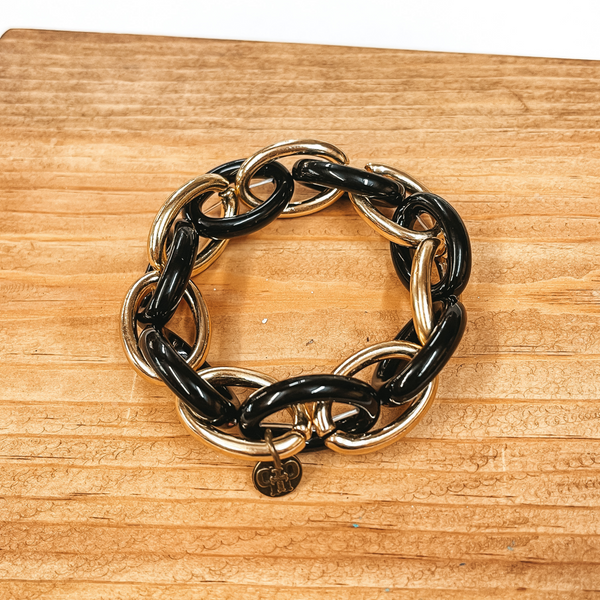 A two toned chained chunky bracelet in gold and black with a Pink Panache pendant. Taken on a brown block and white background.