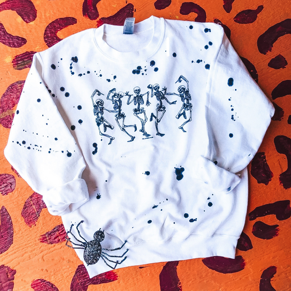 A white sweatshirt with 5 dancing skeletons. Black splattered paint across all of sweatshirt. There is a decorative glittered spider presented in the picture with a pink and yellow leopard background.