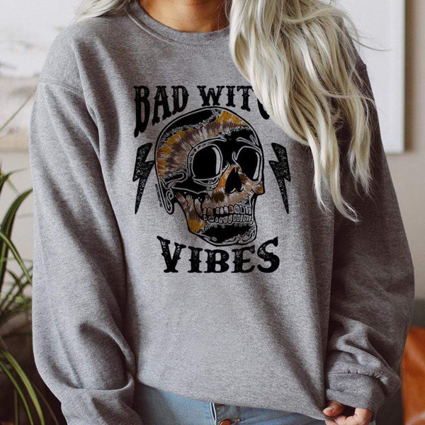 model is wearing a grey sweatshirt which reads "bad witch vibes" in black font with a multi colored skull and lightning bolts. 