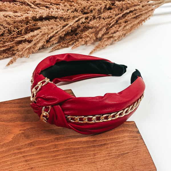 This is a red headband that has a knot in the middle of the headband. There is also a single gold chain that goes through the middle of the headband going through the knot as well. This headband is pictured laying in a piece of wood with tan floral at the top of the picture on a white background.'
