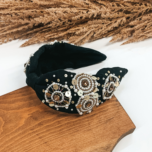 This is a thick, black colored headband that includes white pearl, gold, and bronze detailing. The headband also comes together in the middle with a piece of fabric keeping it small and tied together. This headband is pictured laying in a piece of wood with tan floral at the top of the picture on a white background.