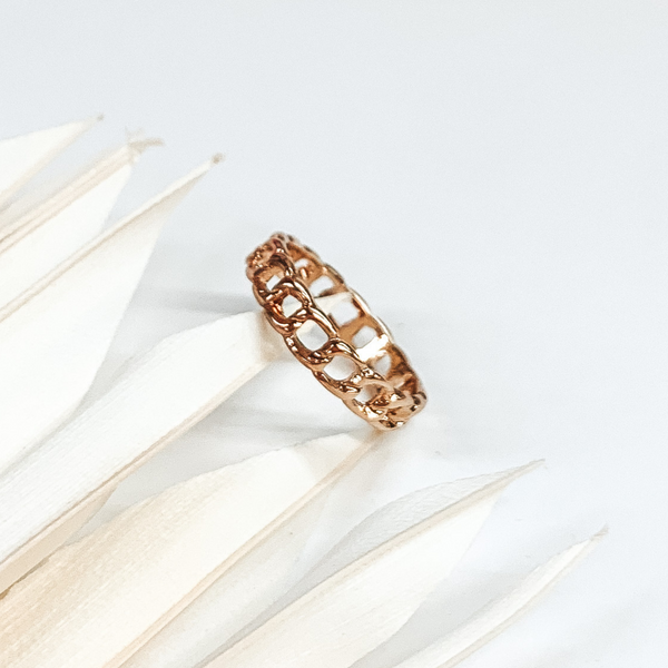 This is a gold chain link ring. this ring is on a white background with some white leaves on the left side of the picture.