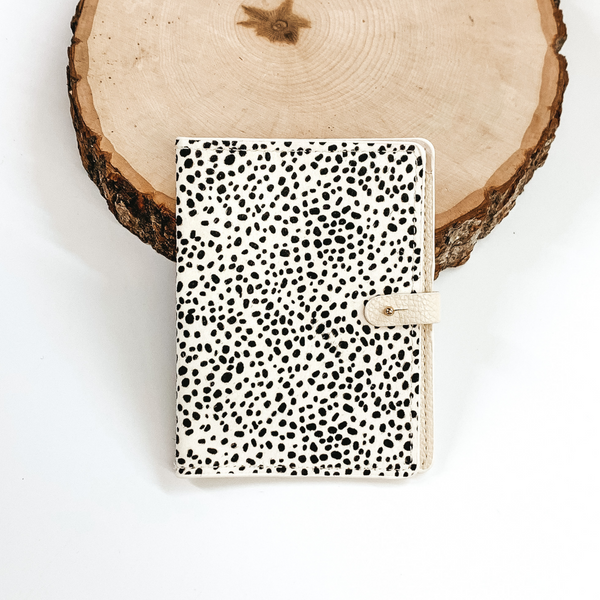 White and black dotted print wallet passport with a gold button to close. This wallet is pictured laying against a piece of wood on a white background.