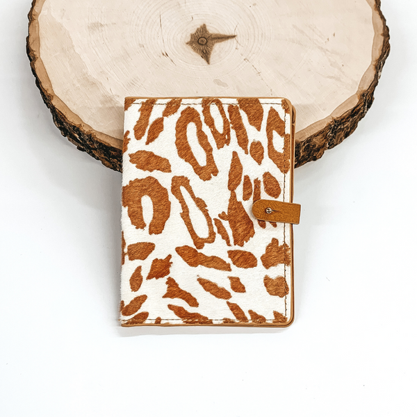 White and tan leopard print wallet passport with a gold button to close. This wallet is pictured laying against a piece of wood on a white background.