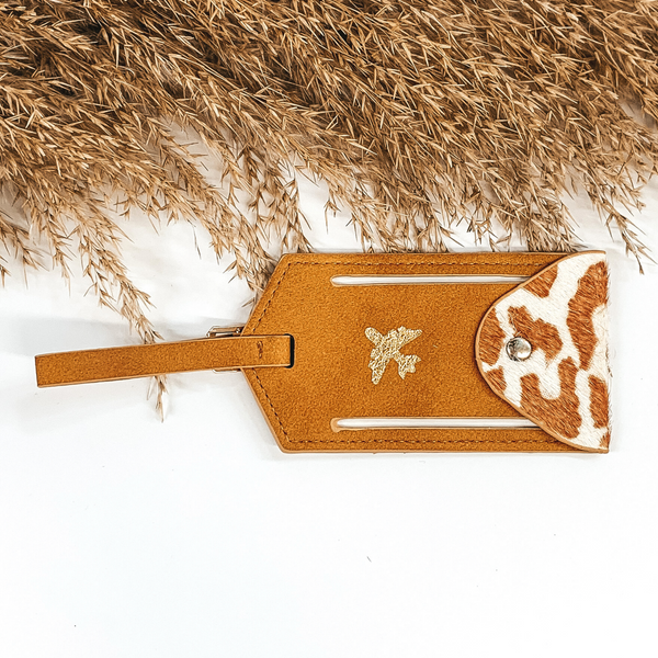 Tan luggage tag with a gold airplane painted on the center. This luggage tag includes a tan strap and an open/close flap that has an ivory and tan leopard print. This is pictured on a white background with light tan floral at the top of the picture. 