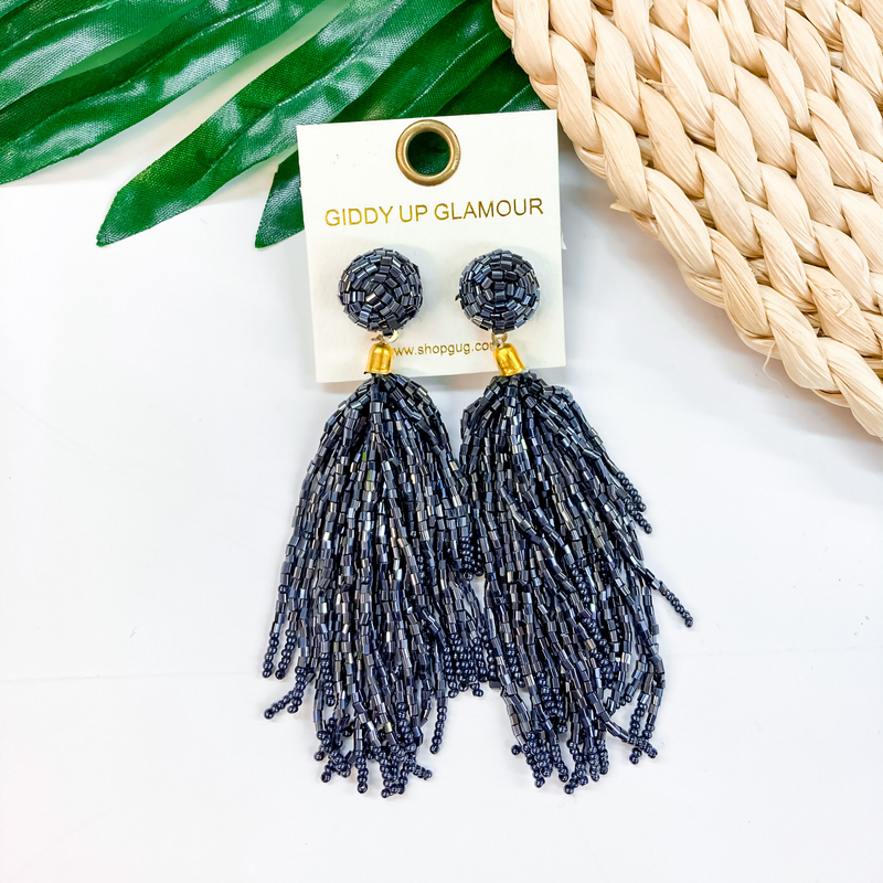 Circle, beaded post back stud earrings with a hanging beaded tassel in black. These earrings are pictured on a white background with a green leaf and tan basket weave in the background.