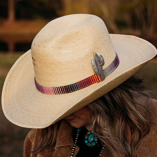 Tan Straw Hat with Curved Brim. Multi-Color Hat Band with Silver Cactus Pin. Model has it paired with a brown jacket and turquoise jewelry. Pictured on wooded background