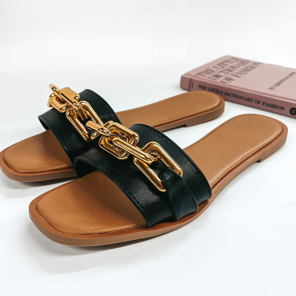 A pair of slide on flat sandals that have a black strap with a gold chain on top. Pictured on white background with fashion book.
