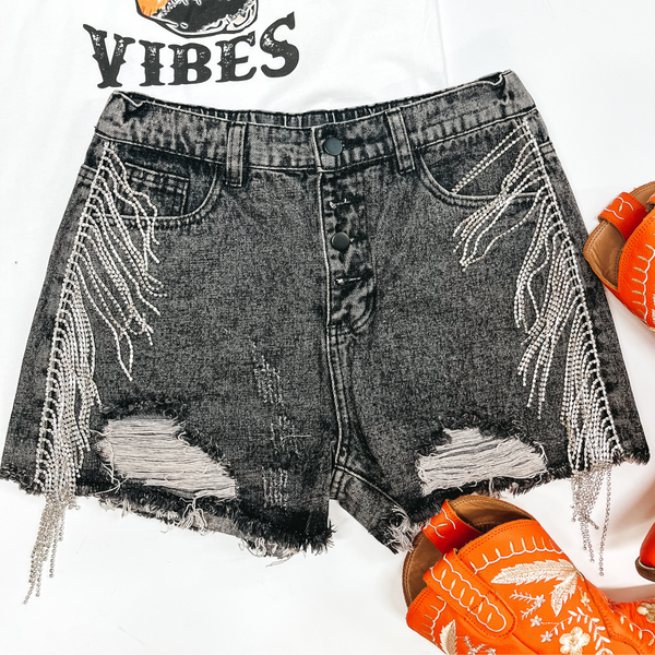 A pair of black wash shorts with a button fly and crystal fringe going down the sides. Pictured on a white background with a white graphic tee shirt and orange boots.