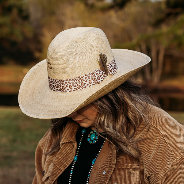 Tan Straw Hat with Curved Brim. Thick Cheetah Bad with Gold Cactus Pin. Model has it paired with a brown jacket and turquoise jewelry. Pictured on wooded background