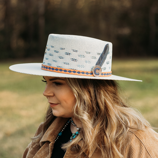 Flat White Brim Straw Hat. Tan and Navy Blue Leather Hat Band with Black Leather Feather. Model has it paired with a brown jacket and turquoise jewelry. Pictured on wooded background