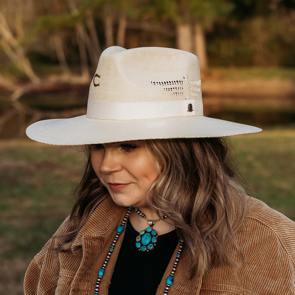 Off White Straw hat with White Silk Hat Band. Model has it paired with a brown jacket and turquoise jewelry. Pictured on wooded background.