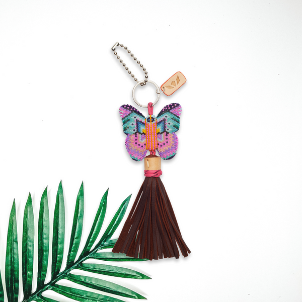 A beaded butterfly charm with a brown leather tassel pictured on a white background with a palm leaf.