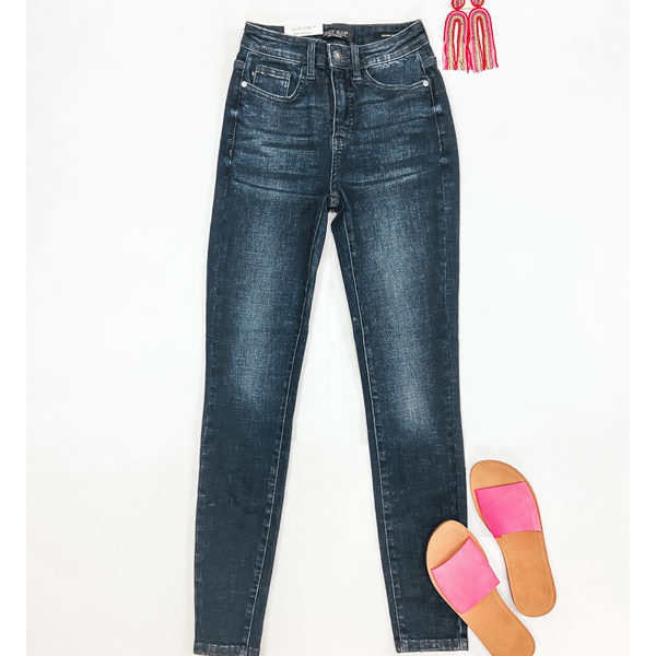 A pair of dark wash high waist skinny jeans pictured on a white background. These jeans are pictured with pink sandals and pink earrings.