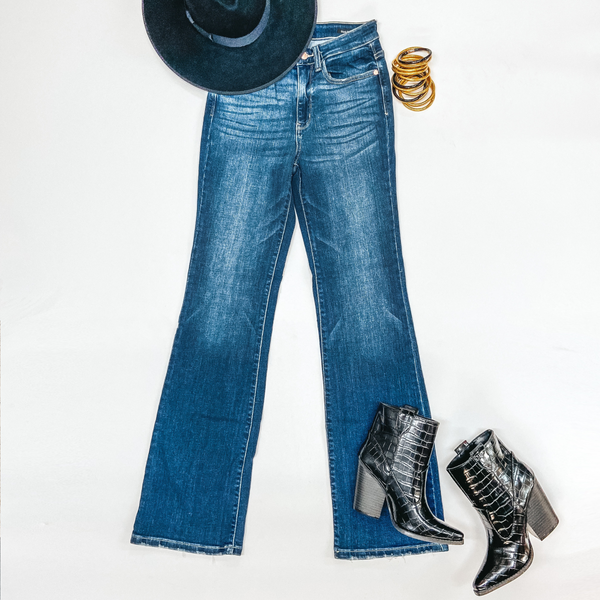 A pair of dark wash bootcut jeans with no distressing. These jeans are pictured on a white background, gold bracelets, a black hat, and black booties.