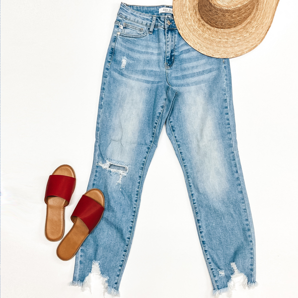 A pair of light wash denim jeans with a destroy knee and hemline. Pictured on a white background with a palm hat and red sandals.