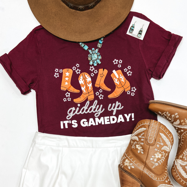 A maroon graphic tee shirt with says "Giddy Up It's Gameday!" in white lettering with orange cowboy boot graphics above the lettering. This tee shirt is pictured on a white background with brown booties, turquoise jewelry. and a brown hat.