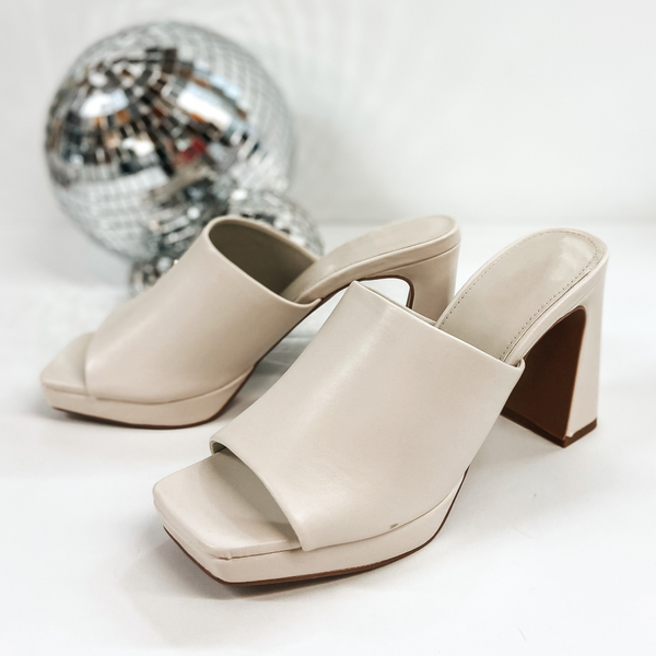 A pair of platform shoes with a slip on upper and open toe. These shoes have a high block heel. The ivory heels are pictured on white background with disco balls.