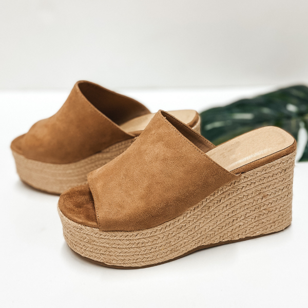 A pair of tan suede slide on wedges that have a rope sole. Pictured on white background with a palm leaf.