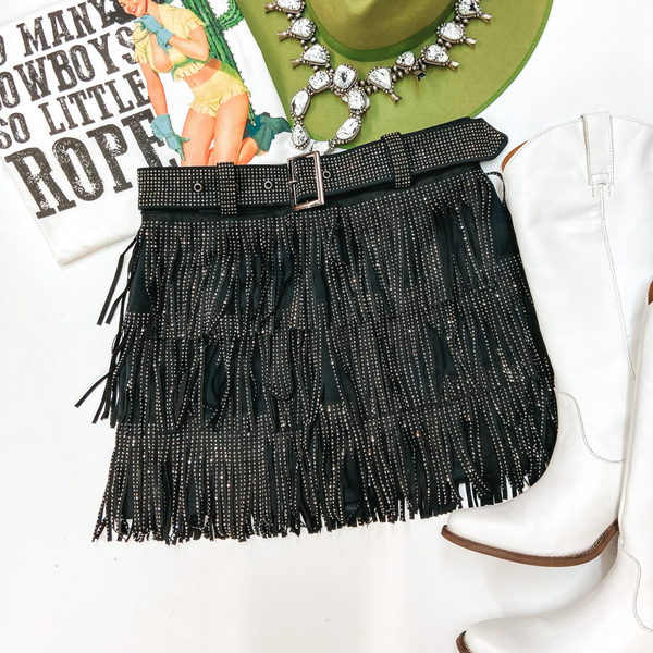 A crystal fringe black skirt with a crystal belt. Pictured with a green hat, white graphic tee, white squash blossom necklace, and white boots.