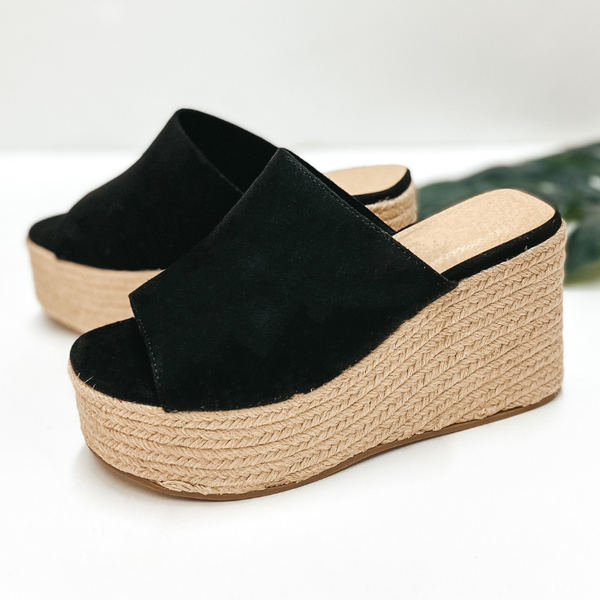 A pair of black suede slide on wedges that have a rope sole. Pictured on white background with a palm leaf.