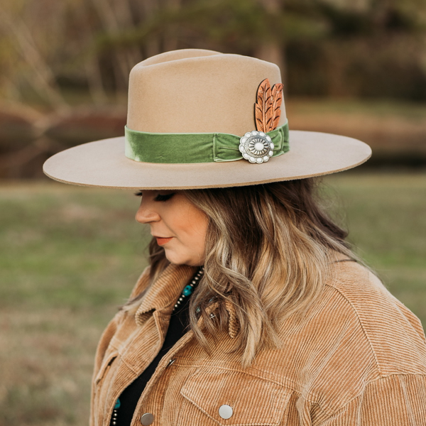 Tan felt hat with a velvet green band, silver concho with ivory stones, and leather feather. Model has it paired with a brown jacket and turquoise jewelry. Pictured on wooded background.