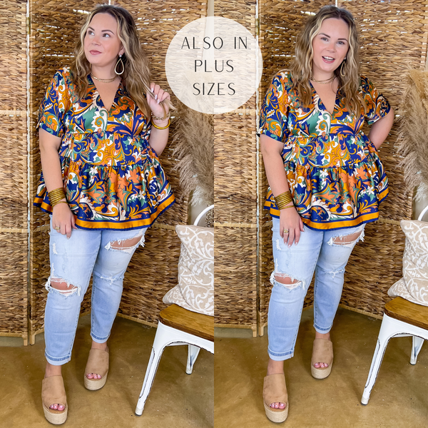 Model is wearing a baroque print top that is a mix of royal blue, gold, and green. This top has short sleeves, a v neckline, and a tiered body. Model has it paired with tan wedges, distressed boyfriend jeans, and gold jewelry.