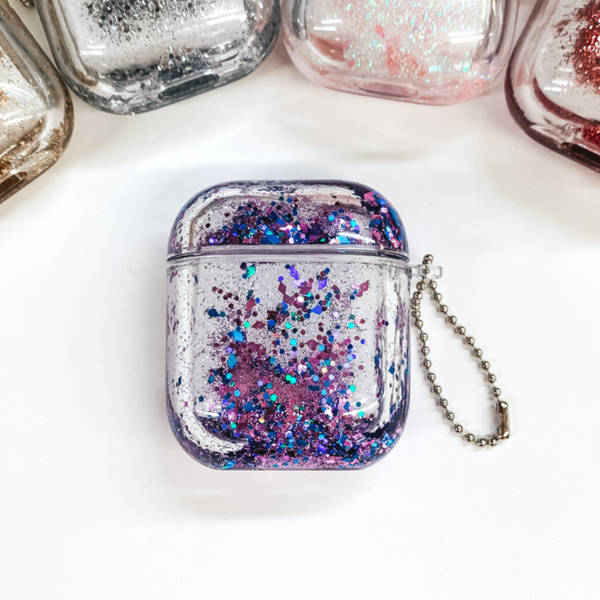 This is a clear airpod case filled with purple, iridescent confetti inside the case. This case has a silver ball chain on one side. This case is pictured on a white background.