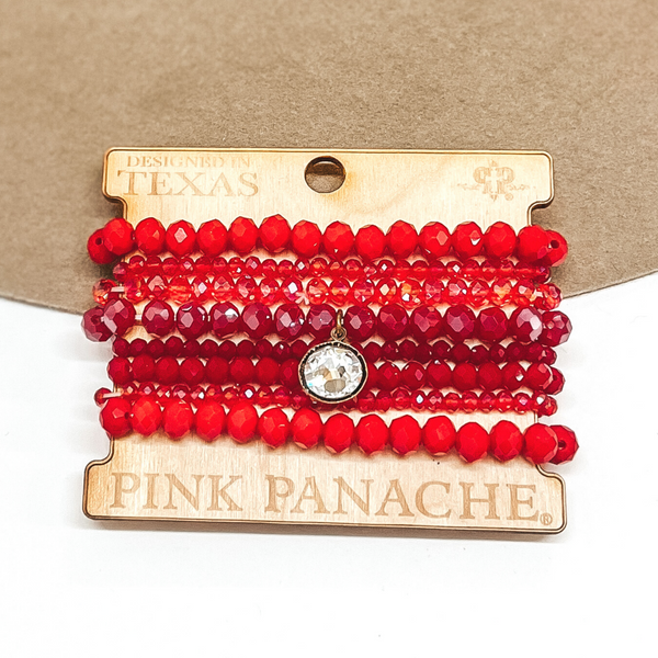 Crystal beaded bracelet set in a mix of reds. One bracelet has a clear circle crystal pendant that has a bronze backing. This bracelet is pictured on a white background in front of a tan hat.