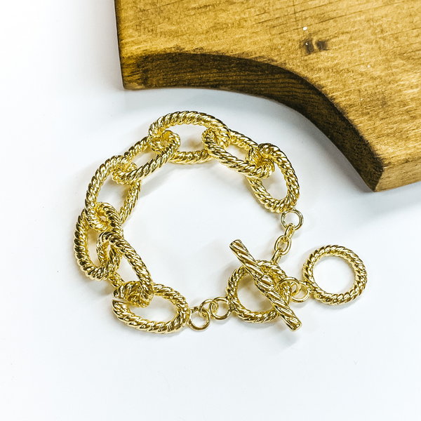 Twisted gold chain bracelet with toggle clasp in gold. This bracelet is pictured on a white background with brown wood in the corner. 