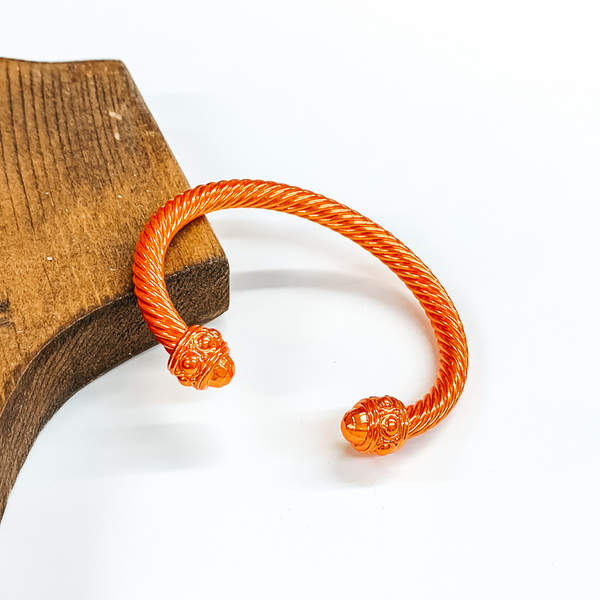 Cable bracelet with cabochon ends in metallic orange. This bracelet is pictured on a white background and leaning on a piece of brown wood. 