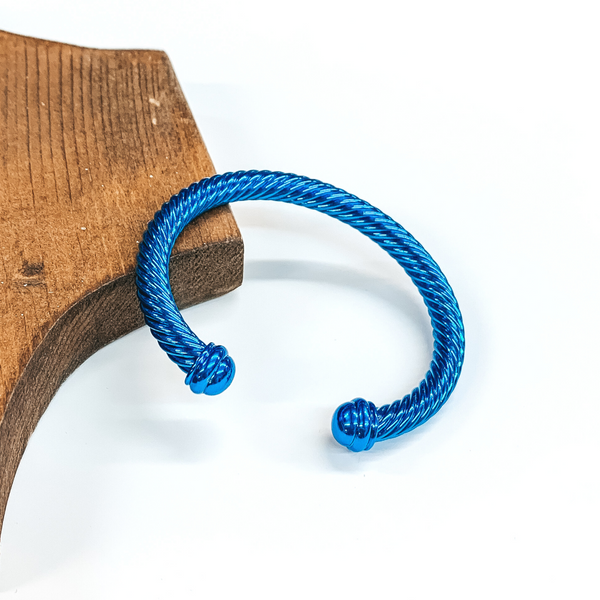 Cable bracelet with cabochon ends in metallic blue. This bracelet is pictured on a white background and leaning on a piece of brown wood.  