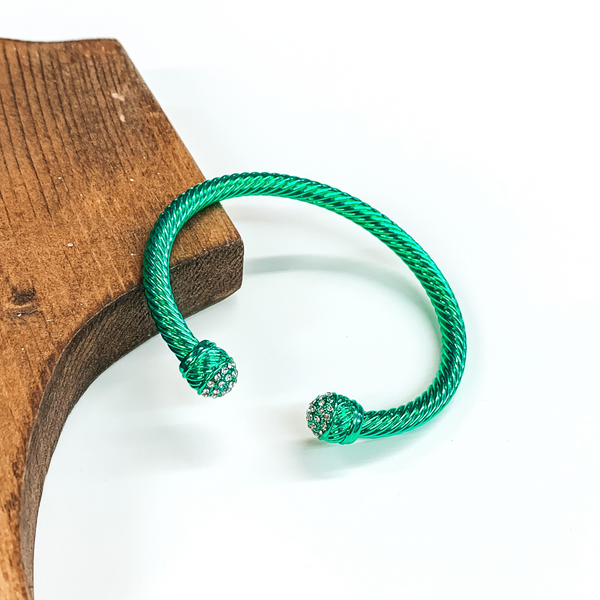 Cable bracelet with clear crystal cabochon ends in metallic green. This bracelet is pictured on a white background and leaning on a piece of brown wood.