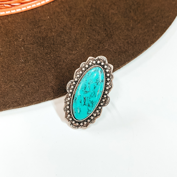 Turquoise oval stone with silver detailing outlining the stone. This ring is pictured on a white and brown background. 