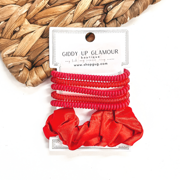 A set of six hair ties, five spiral and one satin scrunchie in red. There are three  solid color spiral hair ties and two transparent. These hair ties are on a white  cardboard piece with a thekitchenapproach logo, they are leaned up against a brown woven  plate and a white background.