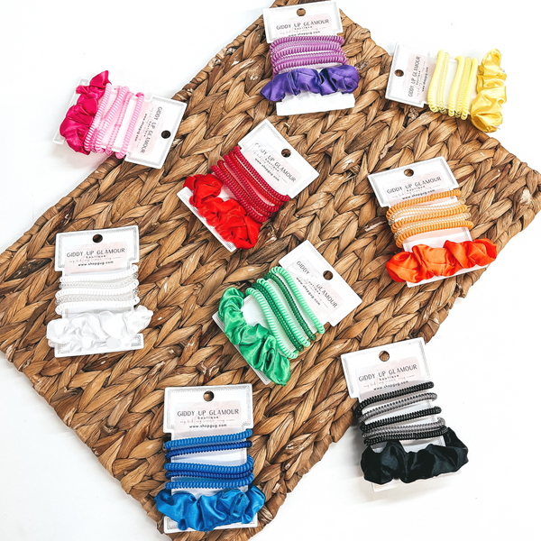 There are nine sets of six spiral and scrunchie hair ties in different colors.  Each set has five spiral hair ties, two transparent and three solid, with one solid  color satin scrunchie. There is in purple, hot pink, yellow, green, orange, red, white, black, and blue. These sets of scrunchies are pictured on a white hair tie  holder. They are laying on a brown woven plate and on a white background.