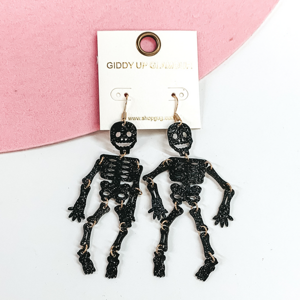 Gold fish hook earrings with a black glitter skeleton with everything collected with gold circles. These earrings are pictured laying partially on a pink hat brim on a white background. 