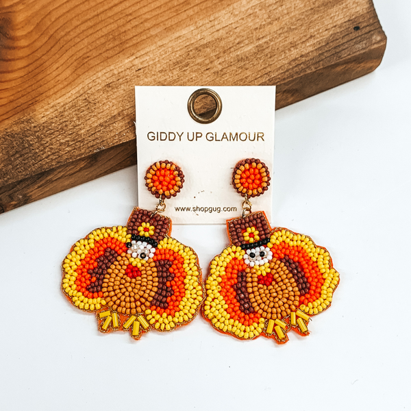 Beaded turkey shaped earrings. These earrings include the colors yellow, tan, bronw, and white. These earrings are pictured in front of a wood block on a white background. 