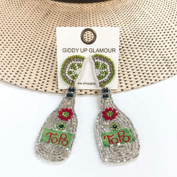 Beaded lime stud earrings with a hanging bottle pendant. The bottle pendant is covered in clear crystals with a red and green beaded flower. It also includes a green stitched area that has the word "ToPo" stitched in red. These earrings are pictured partially laying on a the brim of a straw hat on a white background.