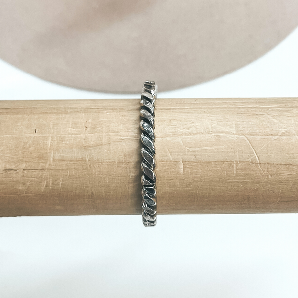 This is a silver rope twist textured bracelet taken on a light brown  bracelet holder and a white background with a beige hat in the back as  decor.