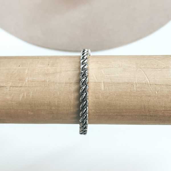 This is a silver rope double twist textured bracelet taken on a light brown  bracelet holder and a white background with a beige hat in the back as  decor.