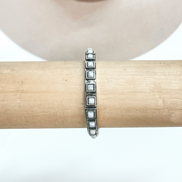 This is a silver bracelet with square white stones all around in a silver  setting. These earrings are taken on a light brown bracelet holder and on  a white background with a beige hat in the back as decor.
