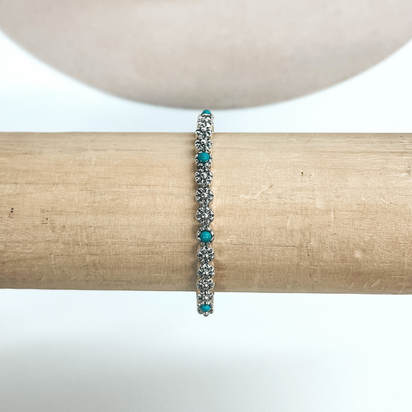 This is a thin silver,flower concho bracelet with small turquoise  stones after every three flowers. These earrings are taken on a light brown bracelet holder and on  a white background with a beige hat in the back as decor.