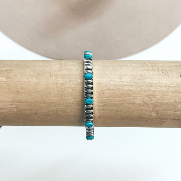 This is a thin silver bracelet, oval engraved texture and small oval  shaped turquoise stones all around. These earrings are taken on a  light brown bracelet holder and on  a white background with a beige hat in the back as decor.